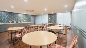 A dining area with round tables and chairs, a kitchen in the background, and a large window providing natural light. - FabHotel Novis Stayz (Noida)