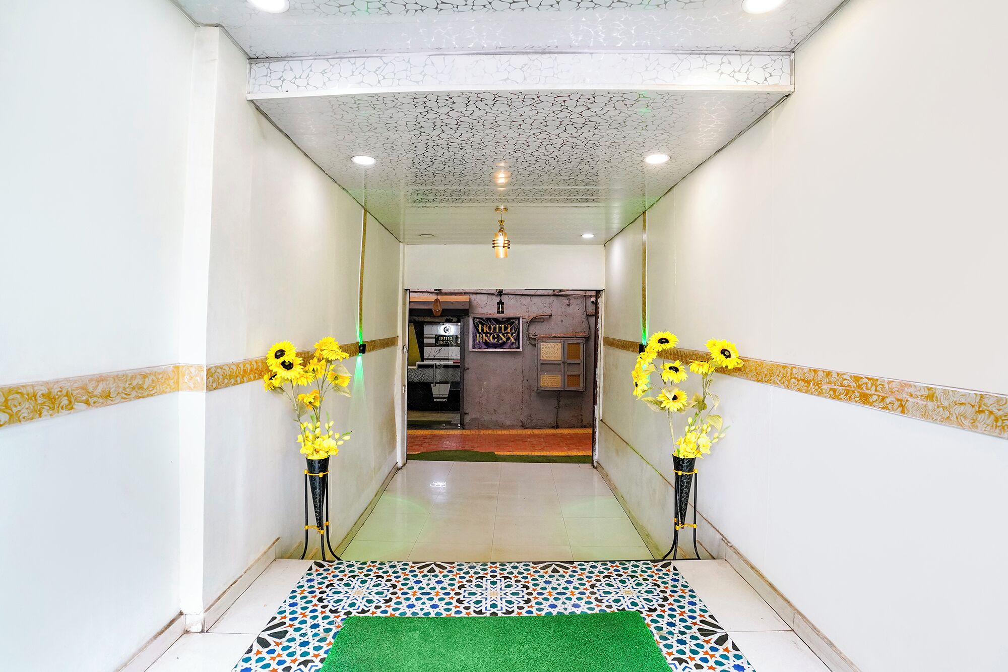 Un pasillo de hotel con baldosas de piso estampadas, alfombra verde y dos jarrones con flores amarillas a cada lado.