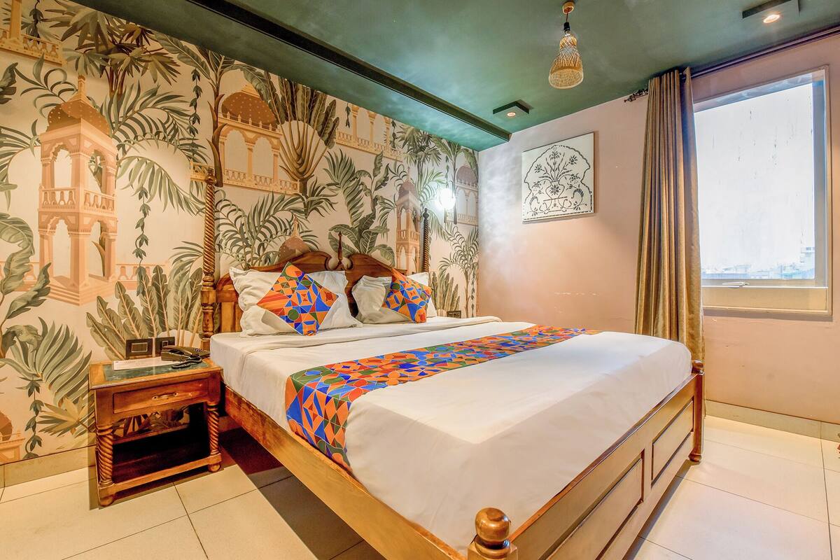 A hotel room with a bed, a nightstand, a wall with a mural, a window with curtains, and a ceiling light.