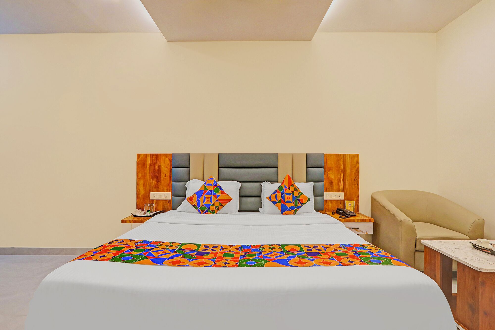 A hotel room with a large bed, a wooden headboard, a beige armchair, and a small wooden side table.