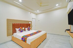 Un dormitorio moderno con cabecera de madera, cama con ropa de cama geométrica de colores, ventilador de techo y televisor de pantalla plana sobre un soporte de madera.