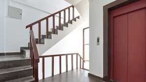 Escada com corrimão de madeira e degraus de azulejos escuros que levam a uma porta vermelha.