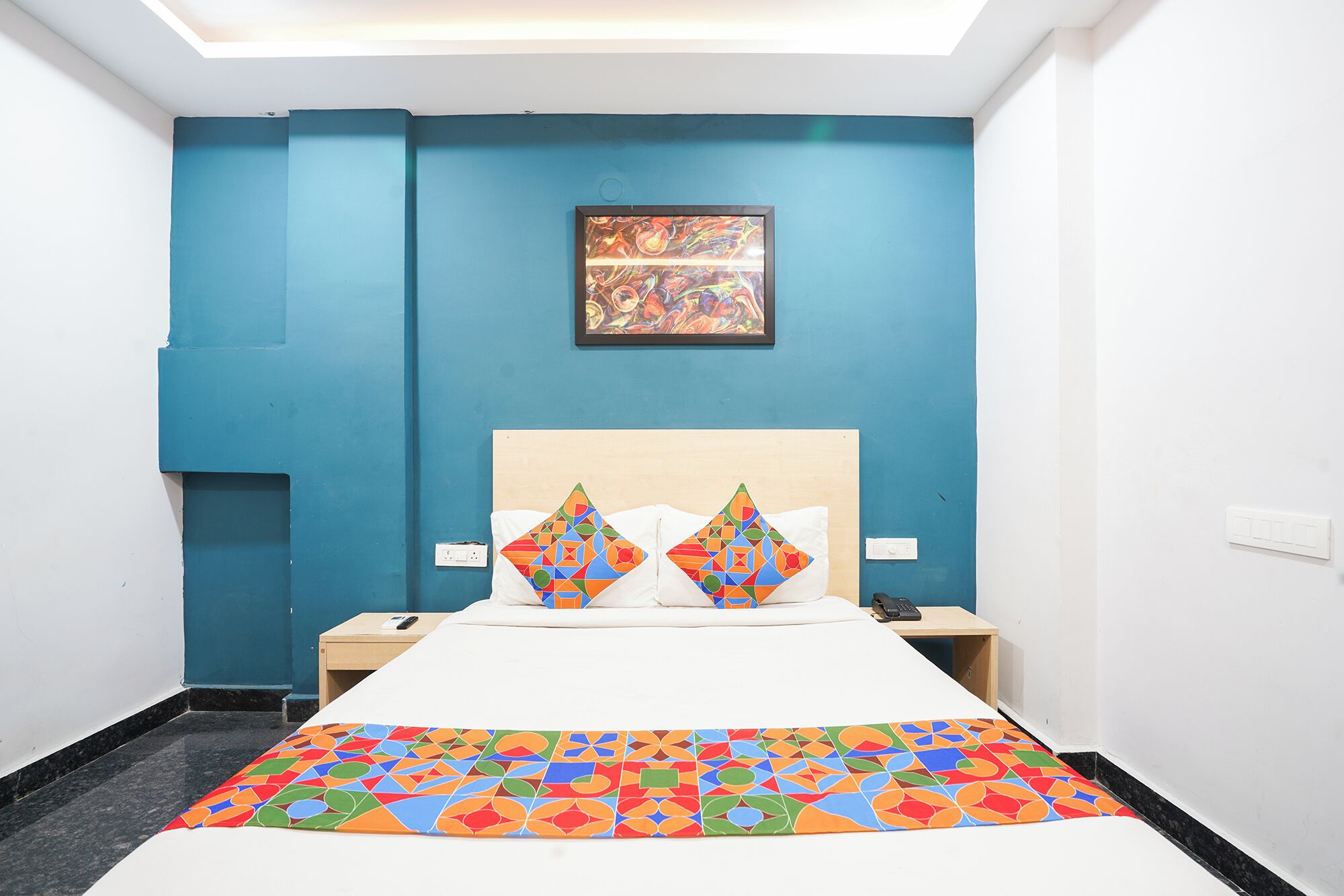 A hotel room with a large bed, two pillows, a colorful bedspread, a painting on the wall, and a nightstand with a phone.