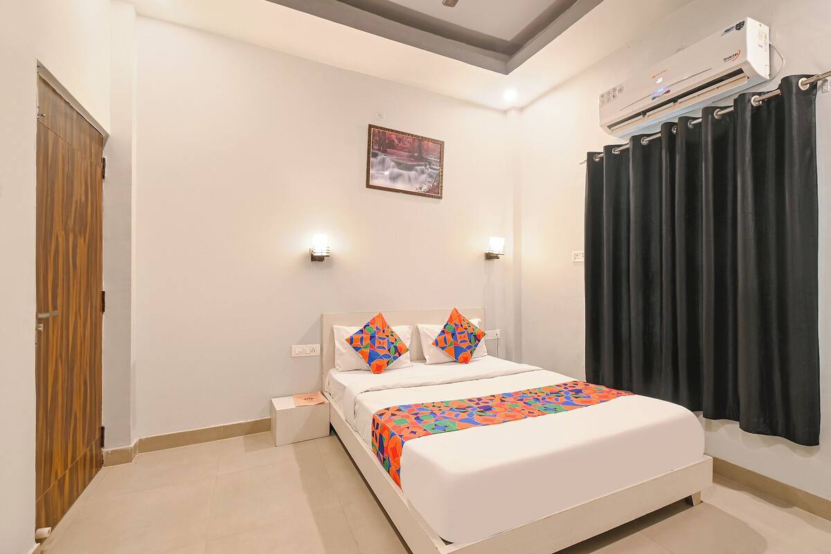 A hotel room with a bed, a wooden door, an air conditioning unit, and a black curtain.