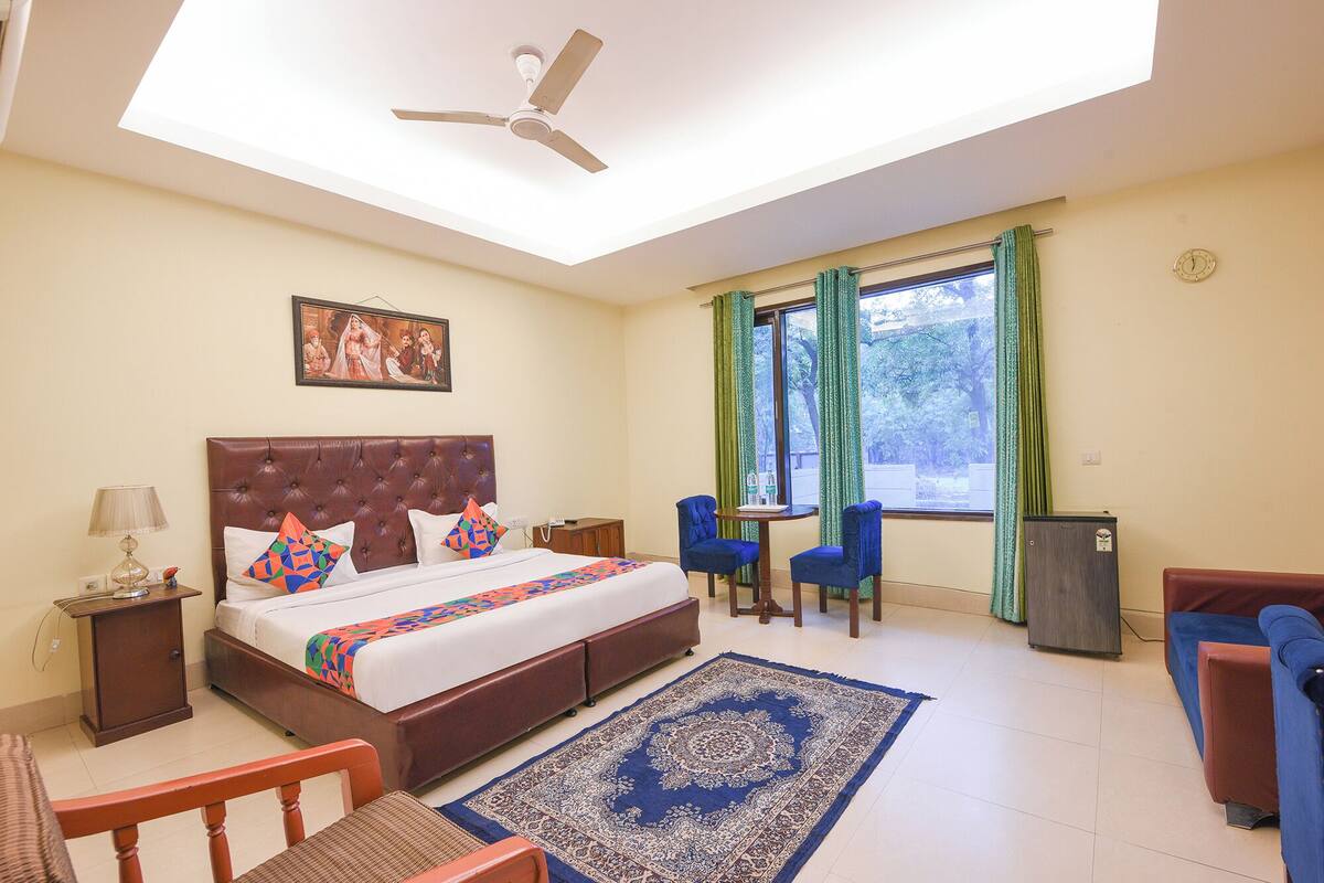A hotel room with a bed, a desk, two chairs, a ceiling fan, a window with curtains, a wall clock, and a small cabinet.