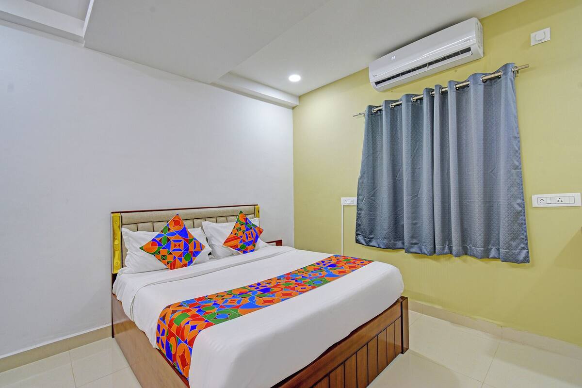 A hotel room with a bed, a window with curtains, an air conditioning unit, and a wall socket.