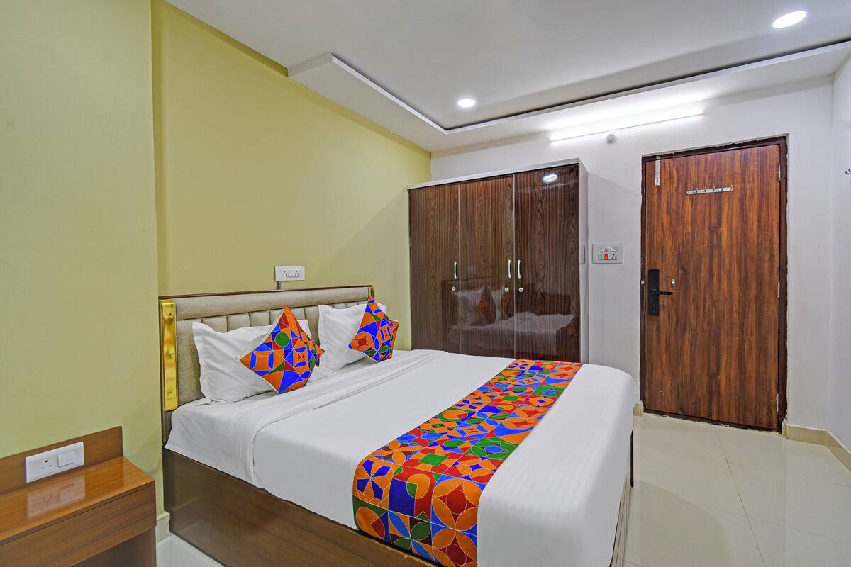 A neatly made bed with a colorful geometric-patterned bedspread and pillows, a wooden wardrobe, and a wooden nightstand with a power outlet.