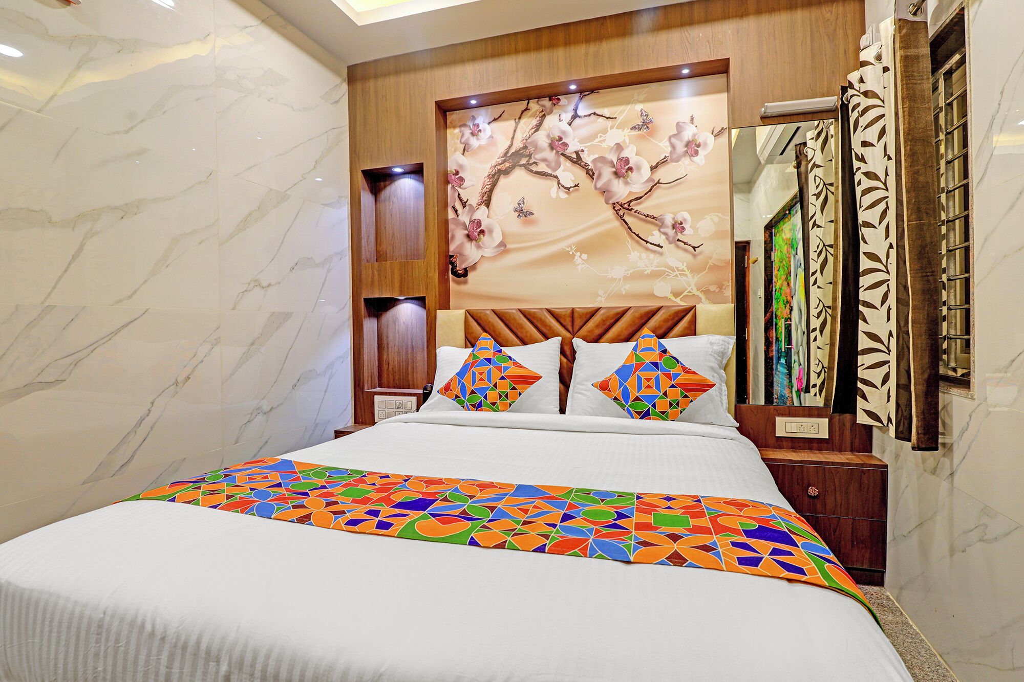 A hotel room with a marble wall, a bed with colorful pillows, a wooden headboard, and a large mirror.