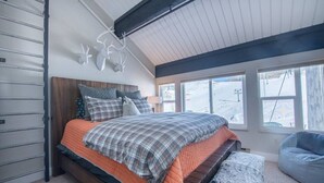 A bedroom with a bed, a bench, a wall-mounted light fixture, and a window with a view of a snowy landscape. - Mountain-front Condo Sleeps 8 + Sauna (Crested Butte)