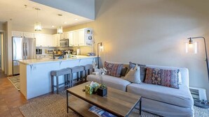 A modern kitchen with white cabinets, a central island, and bar stools. There is a living area with a sofa and a coffee table. - Mountain-front Condo Sleeps 8 + Sauna (Crested Butte)