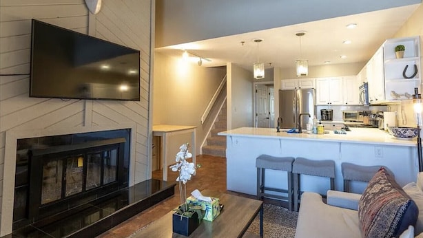 A modern living room with a fireplace, a flat-screen TV mounted on the wall, and a kitchen area with a central island and bar stools. - Mountain-front Condo Sleeps 8 + Sauna (Crested Butte)