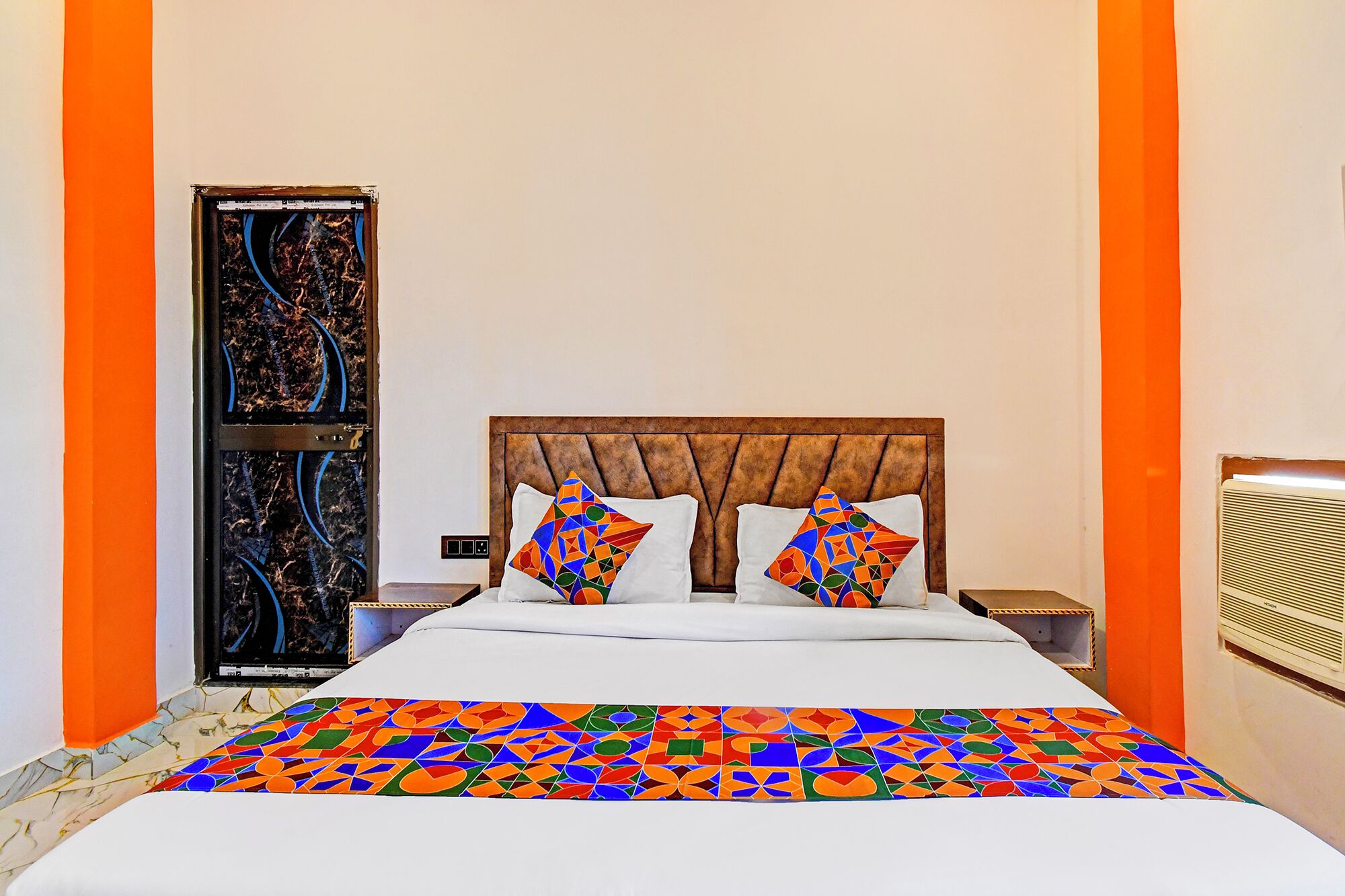 Ett hotellrum med en sänggavel i trä, en säng med färgglatt sänglinne med geometriska mönster och orange accentväggar.