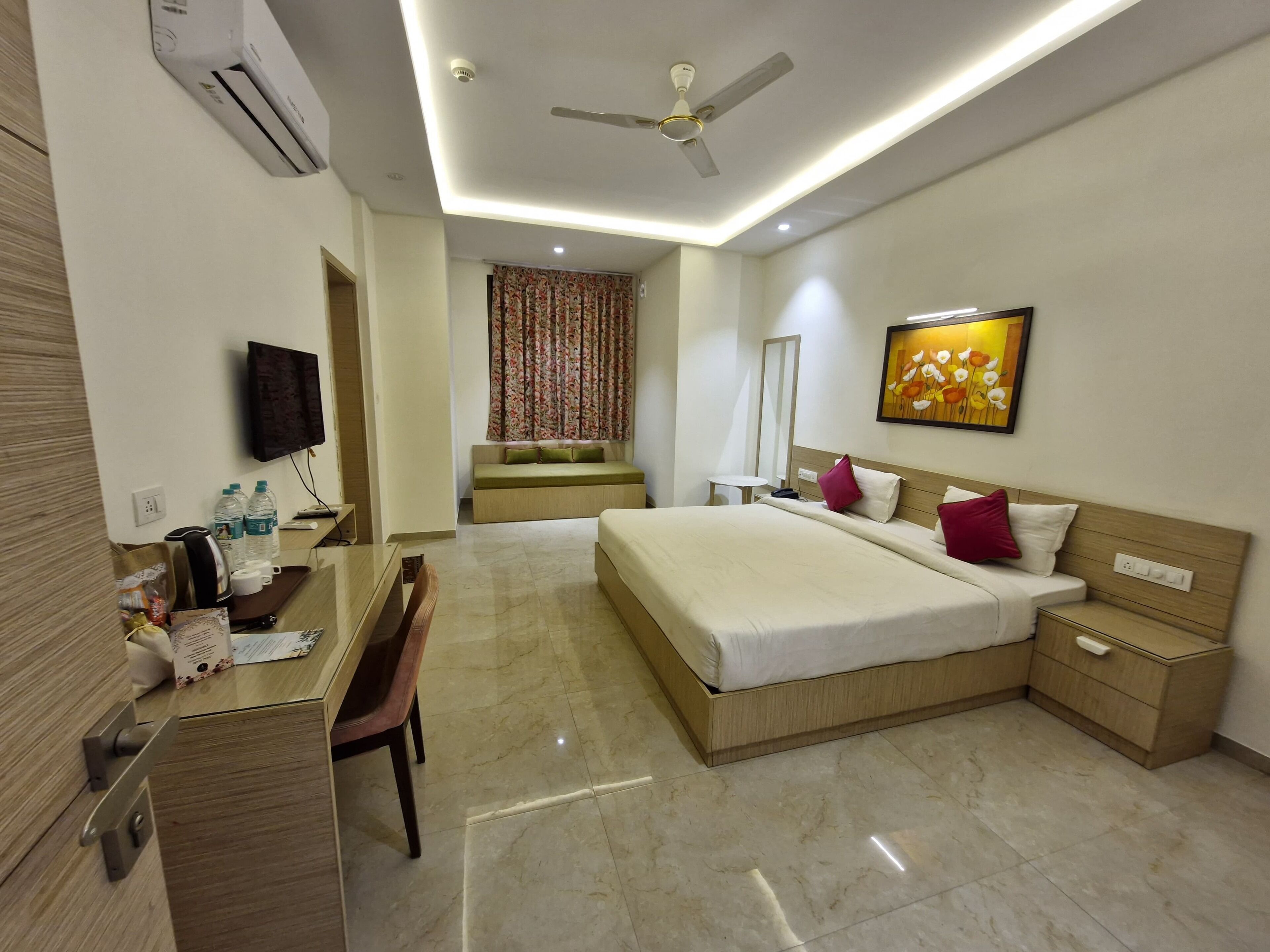 A hotel room with a bed, bedside tables, a desk with a chair, a flat-screen TV, a ceiling fan, and a painting on the wall.