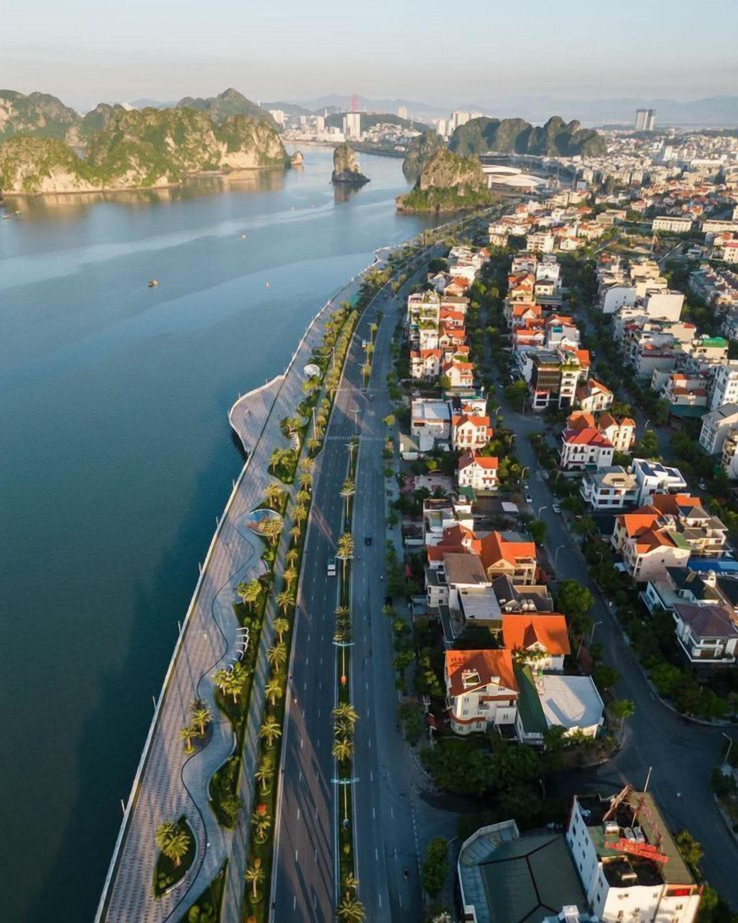 A coastal cityscape with a prominent waterfront, a curved road lined with palm trees, and residential buildings with red roofs.