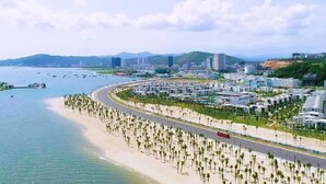 Un paysage urbain côtier avec une plage, une route sinueuse bordée de palmiers et des bâtiments modernes.