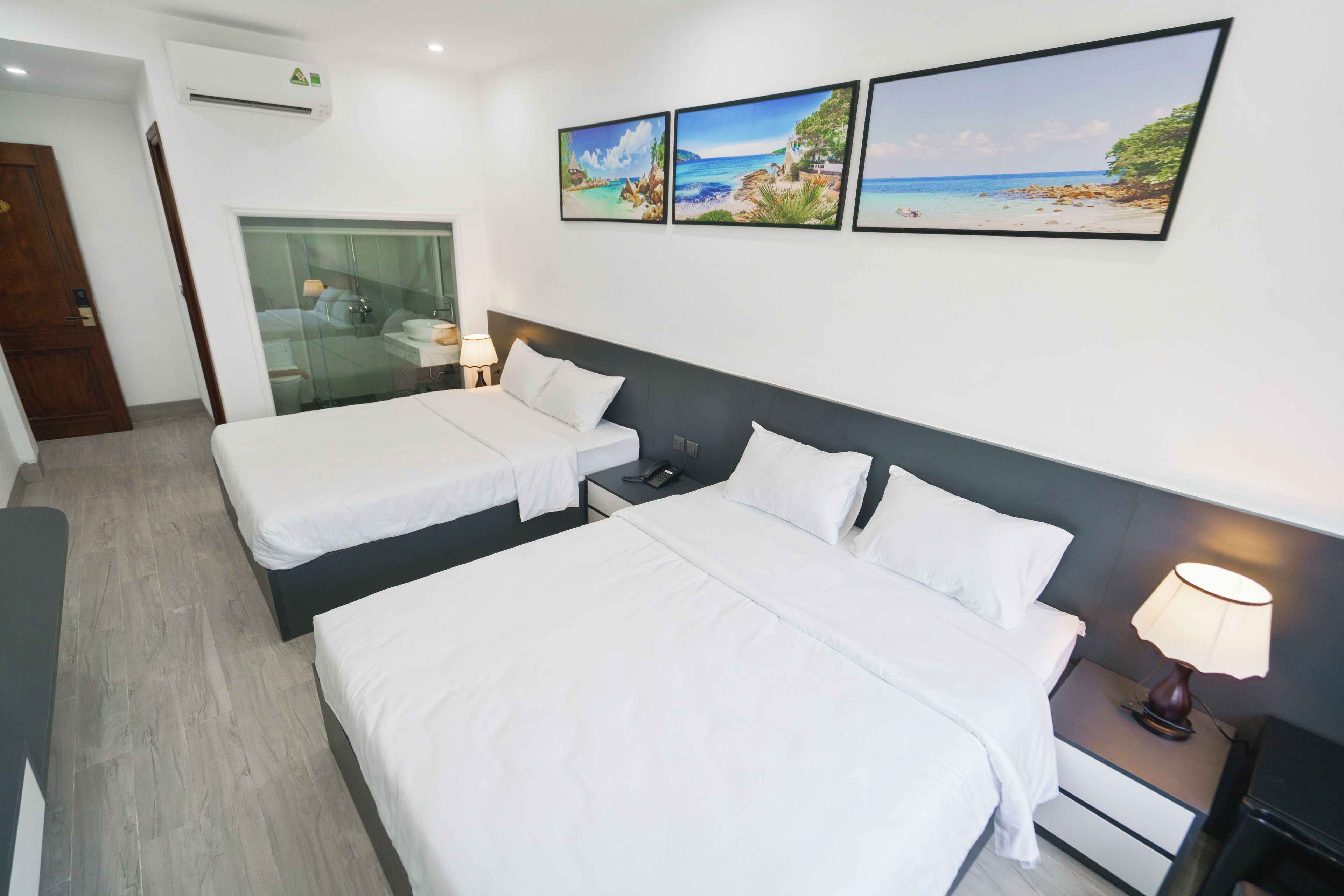 Habitación de hotel con dos camas individuales, una mesita de noche con lámpara, cabina de ducha con paredes de vidrio y tres cuadros enmarcados en la pared.