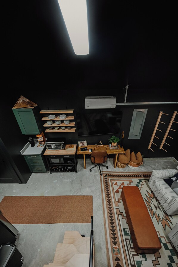 Una cocina compacta con microondas, tostadora y cafetera, un pequeño comedor con una silla y mesa, y una alfombra estampada.