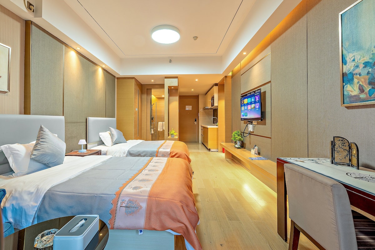 Quarto de hotel com duas camas, uma escrivaninha, televisão e banheiro visível por uma porta aberta.