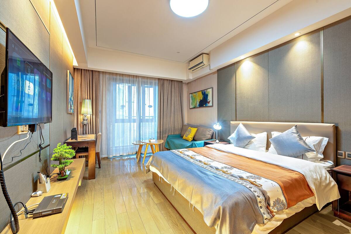 Quarto de hotel moderno com uma cama grande, uma escrivaninha com computador, uma televisão e uma janela com cortinas.
