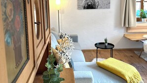 A living room with a grey sofa, a wooden cabinet with a framed picture, a wall-mounted lamp, a black and white bull painting, and a small round table with a plant. - Apartment Liesl 75sqm for max. 5 persons (Schechen)