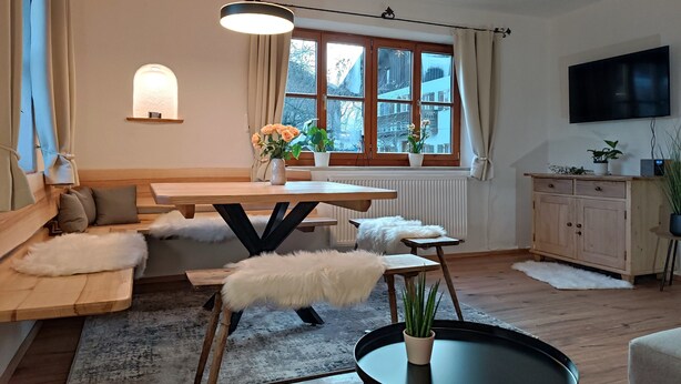 A modern living room with a wooden dining table, a sofa, a white furry ottoman, a round black table, and a TV mounted on the wall. - Apartment Liesl 75sqm for max. 5 persons (Schechen)