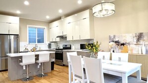 A modern kitchen with white cabinets, a central island with bar stools, a dining table with chairs, and a hanging light fixture.
