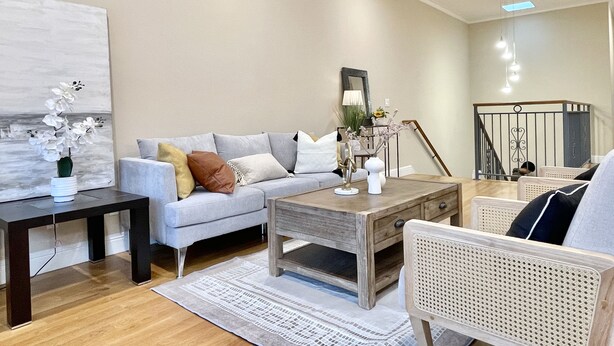 A living room with a grey sofa, a wooden coffee table, a white vase with flowers, and a staircase with a railing.