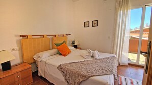 A neatly made bed with a blanket, pillows, and a folded towel. There is a nightstand with a lamp and a small table with a vase of flowers. The room has a window with curtains and a view of buildings outside. - El Alfar - Denia, Costa Blanca (Denia)