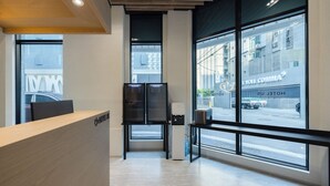 Zona de recepción de un hotel con un mostrador de madera, una pantalla digital y amplios ventanales que dan a la calle y otros edificios.