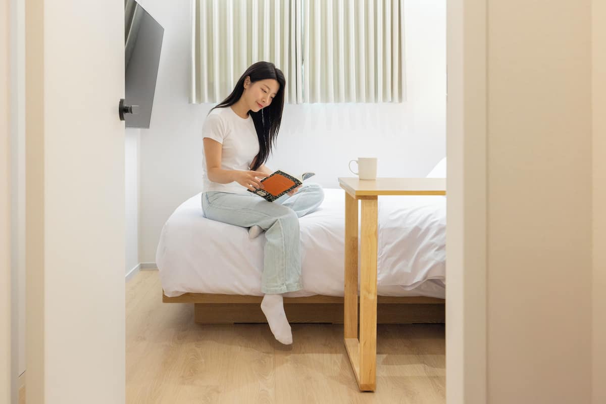 Eine Person sitzt auf einem Bett in einem Zimmer mit Holzboden, einer weißen Wand und einem Fenster mit vertikalen Jalousien.
