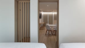 Habitación de hotel con dos camas, un separador de madera, una zona de comedor con mesa y sillas, y vista de la habitación contigua.