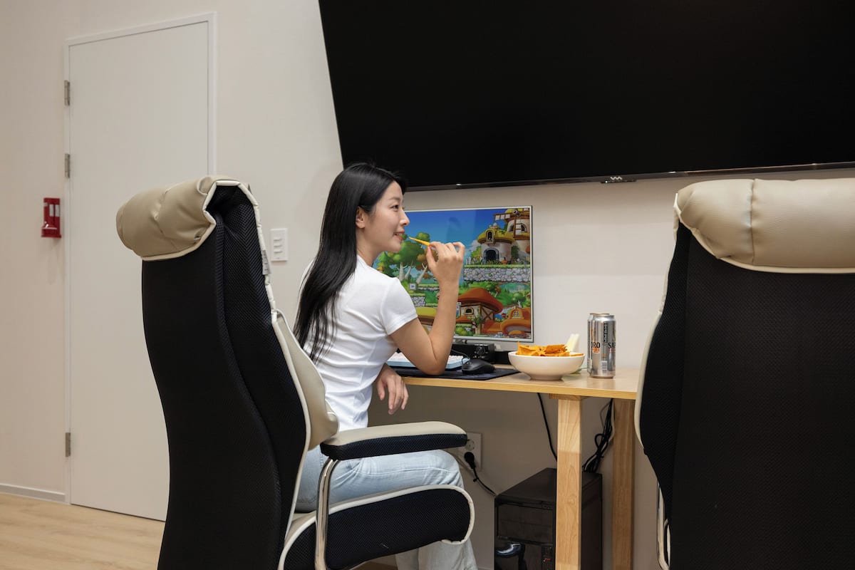 Eine Person sitzt an einem Schreibtisch mit einem Computermonitor, einer Schüssel mit Essen und einer Getränkedose. Zwei Stühle mit Kopfstützen, eine Tür und ein an der Wand befestigter Fernseher sind ebenfalls vorhanden.