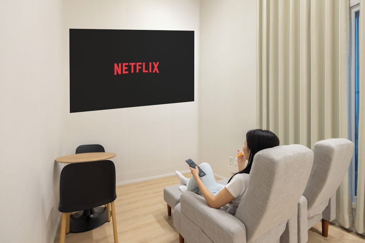 Eine Person sitzt auf einem grauen Sofa in einem Raum mit einem schwarzen Bildschirm, auf dem 'NETFLIX' angezeigt wird, und einem kleinen Tisch mit einem Stuhl.