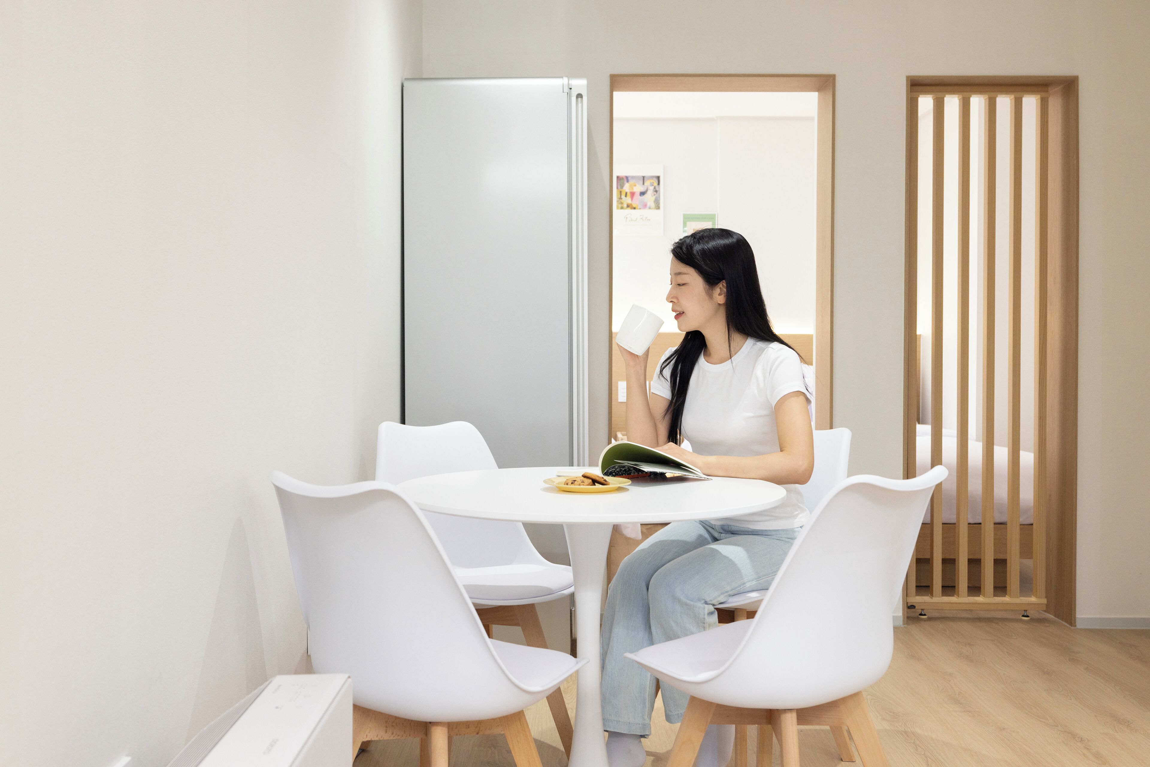 A woman sitting at a white table with a book and a cup, in a room with a white chair, a wooden floor, and a white wall.