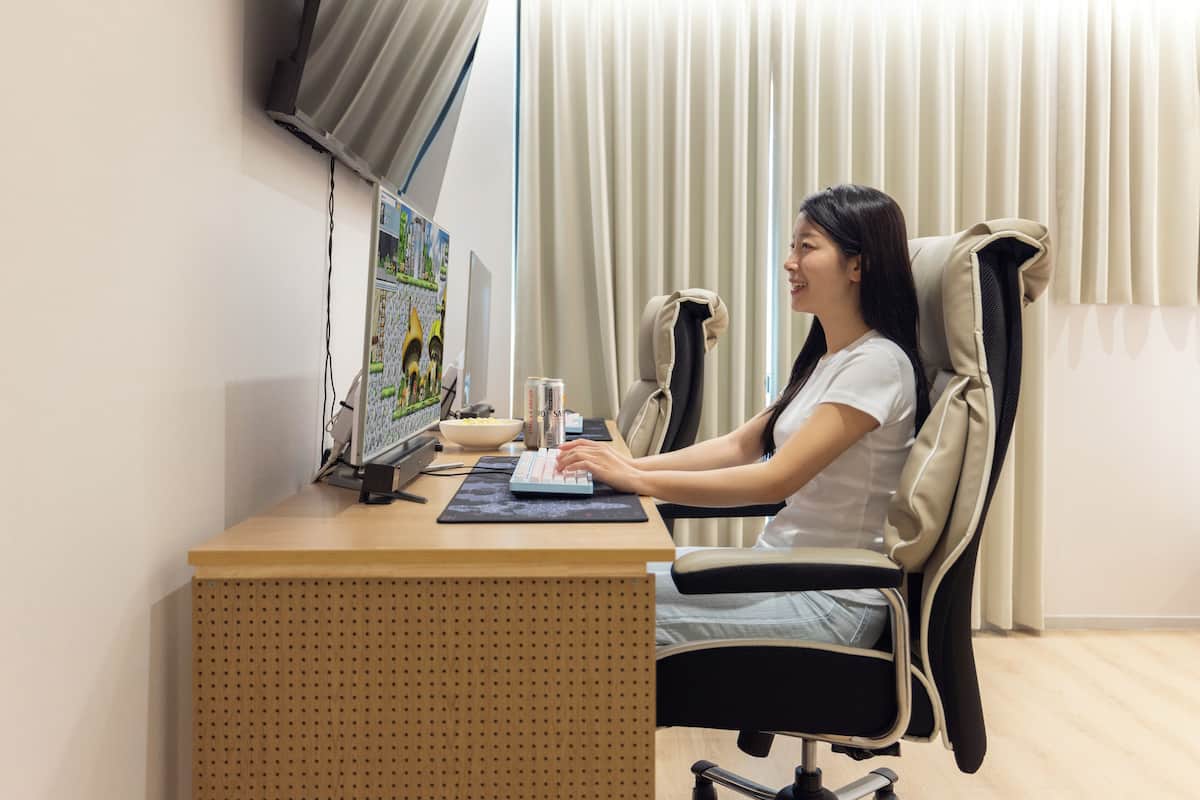 Eine Person sitzt an einem Schreibtisch mit einem Computer, einer Tastatur und einer Maus. Ein Stuhl mit hoher Rückenlehne und Sitzkissen ist vorhanden. Im Hintergrund ist ein Fenster mit Vorhängen zu sehen.