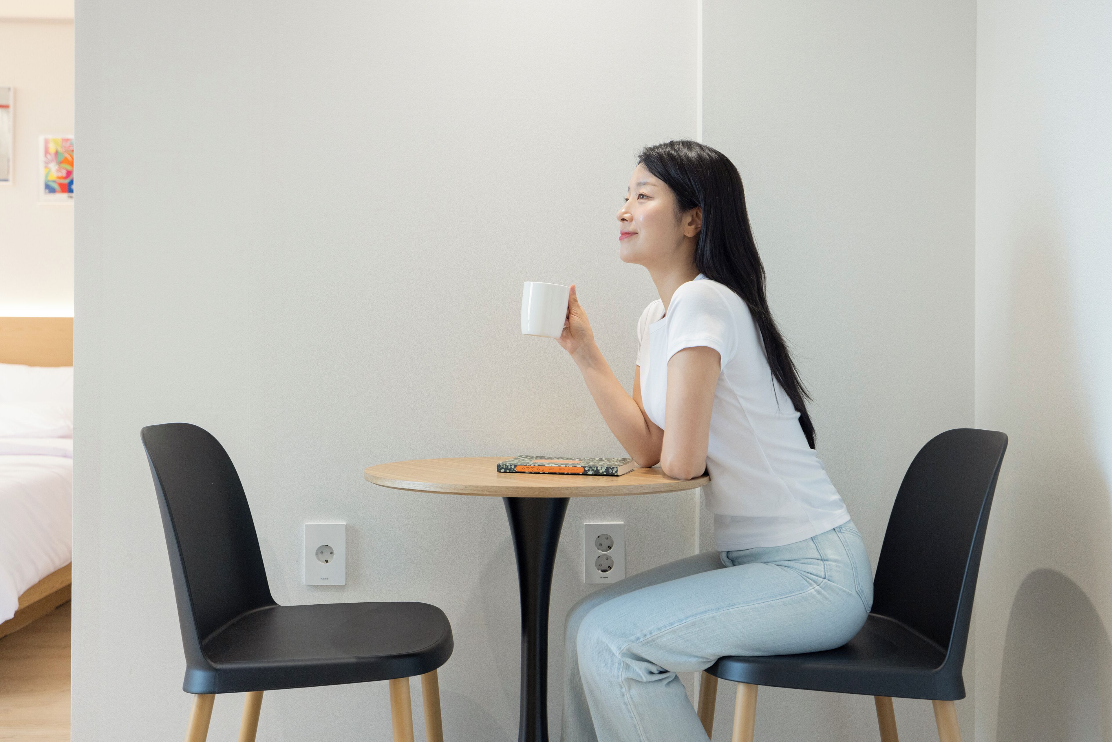 A person sitting at a small round table with a book, holding a white mug. There are two black chairs, one on each side of the table. The room has a simple, clean design with a white wall and a bed in the background.
