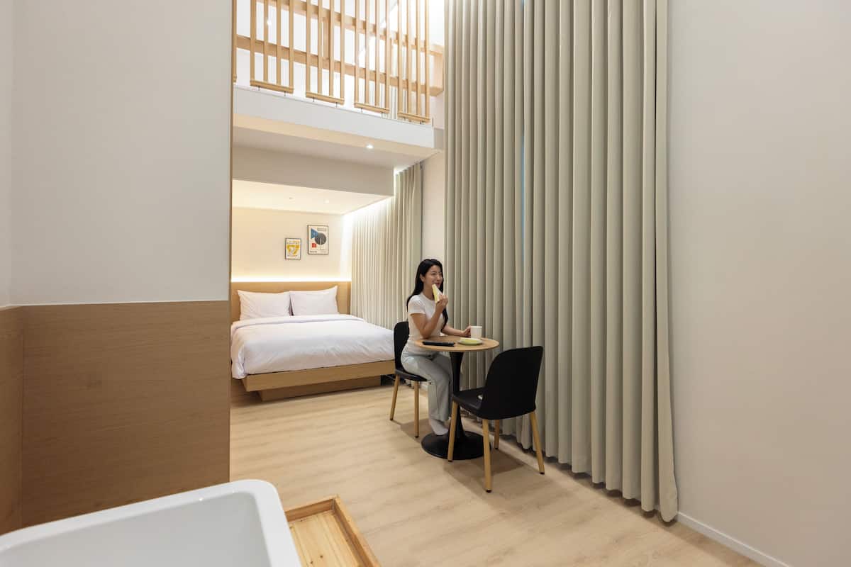 Ein modernes Hotelzimmer mit einem Bett, einem Esstisch und einem Stuhl. Ein Fenster mit Vorhängen und eine Holzbalkendecke sind ebenfalls vorhanden.