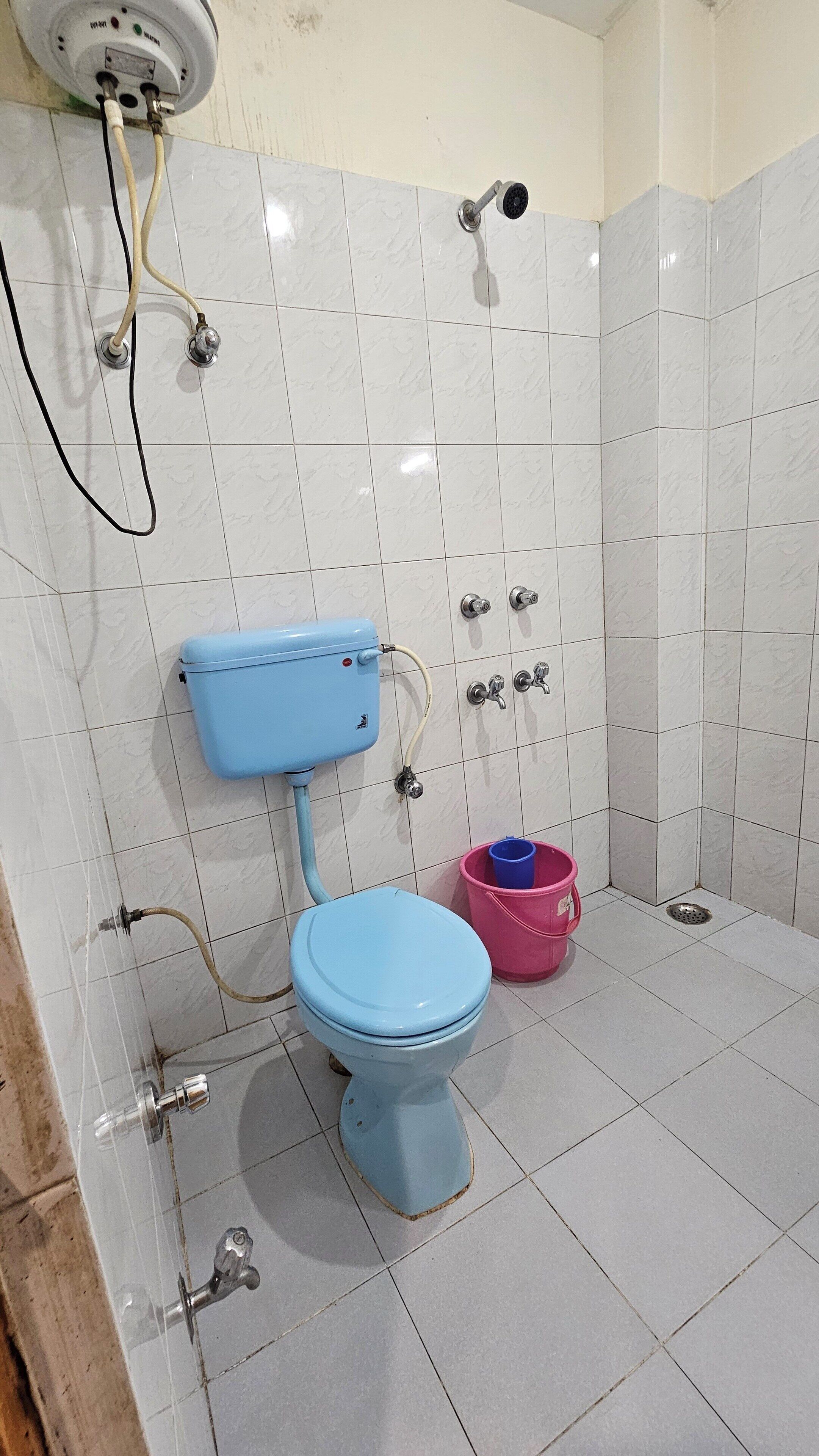 Casa de banho com sanita azul, balde rosa e chuveiro.