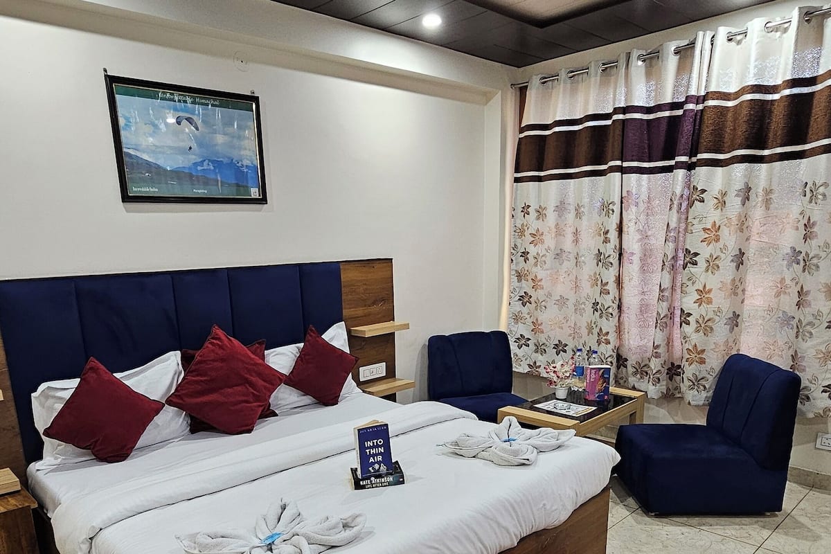 A hotel room with a large bed, a ceiling fan, a window with curtains, a wooden nightstand, and a red patterned rug.