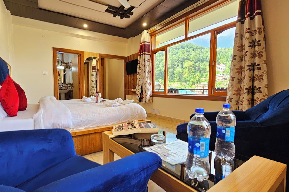 A hotel room with a bed, two blue sofas, a wooden table with water bottles, and a large window with a view of greenery.