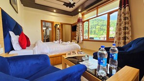 A hotel room with a bed, two blue sofas, a wooden table with water bottles, and a large window with a view of greenery. - Hotel Alpine Vista (Dharamshala)