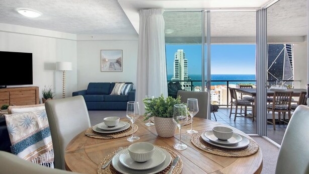 A dining area with a view of the ocean and buildings, a living room with a sofa and TV, and a balcony with a table and chairs. - Solnamara Apartments (Burleigh Heads)
