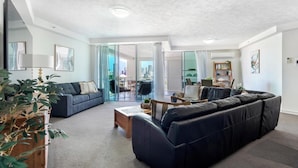 A spacious living room with a dark leather sofa, a grey sofa, and a wooden coffee table. There is a bookshelf and a large window with a view of the city. - Solnamara Apartments (Burleigh Heads)