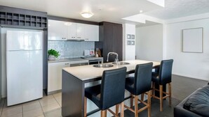 A modern kitchen with a central island, bar stools, and a refrigerator. - Solnamara Apartments (Burleigh Heads)