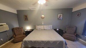 Ein Schlafzimmer mit einem großen Bett, zwei Stühlen, einem Nachttisch, einer Wanduhr und einem Deckenventilator.