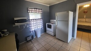 Eine kompakte Küche mit Mikrowelle, Herd und Kühlschrank. Ein Fenster mit karierten Vorhängen, ein Deckenventilator und ein Mülleimer sind ebenfalls vorhanden.