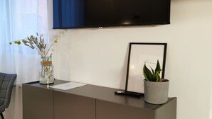 Un salon moderne avec un téléviseur à écran plat fixé au mur, un meuble gris orné d’un vase de fleurs séchées, un tableau encadré et une plante en pot.