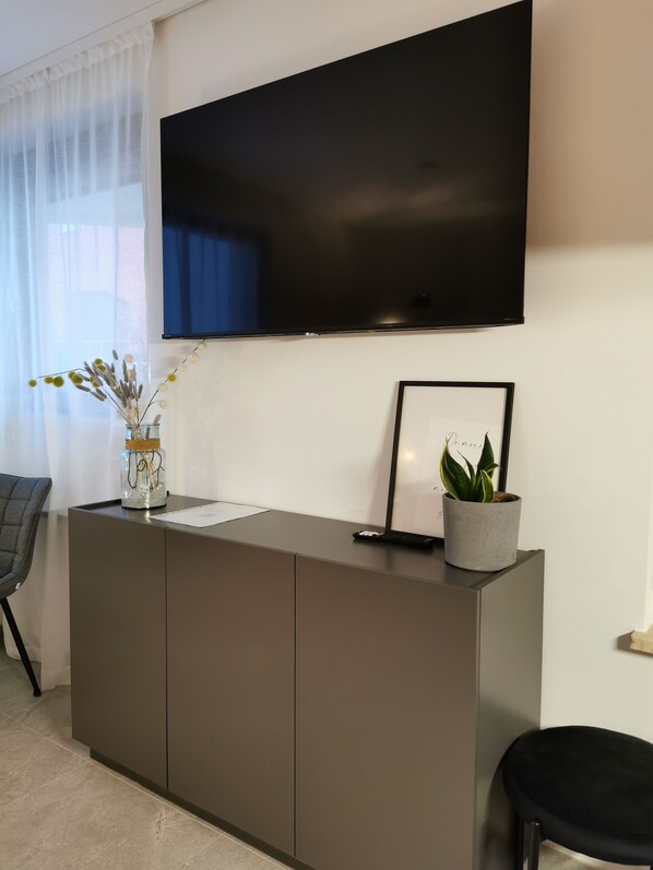 A modern living room with a flat-screen TV mounted on the wall, a grey cabinet with a vase of dried flowers, a framed picture, and a potted plant. - SmartApart - Design Apartment - Favorite Place- Full Equipment- Public Transport (Nürnberg)
