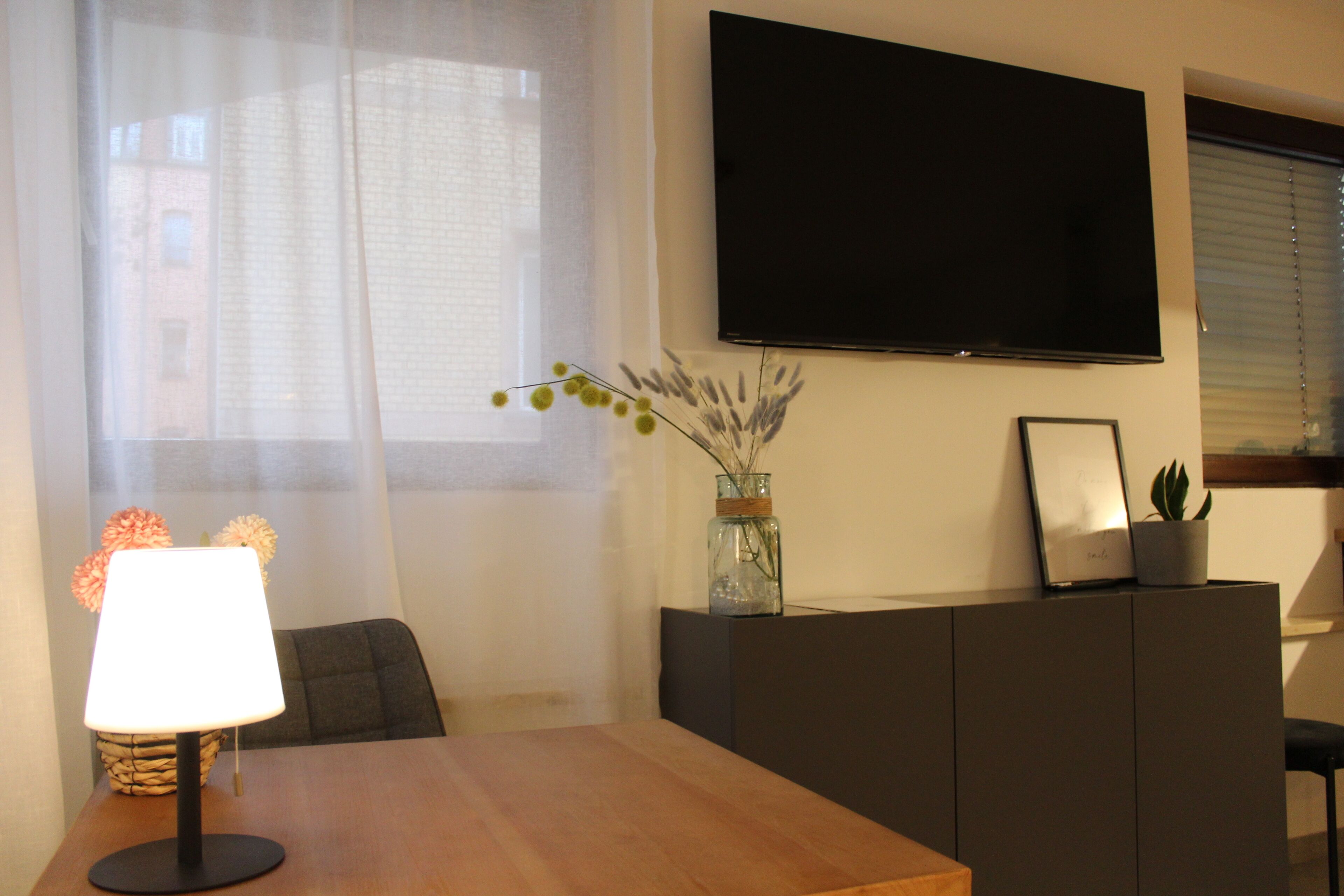 A room with a wooden table, a lamp, a TV mounted on the wall, a vase with dried flowers, and a potted plant.