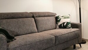 Un salon moderne avec un canapé gris à motifs, un accoudoir noir, un coussin vert et blanc à motifs de feuilles, un lampadaire et une photographie en noir et blanc encadrée accrochée au mur.